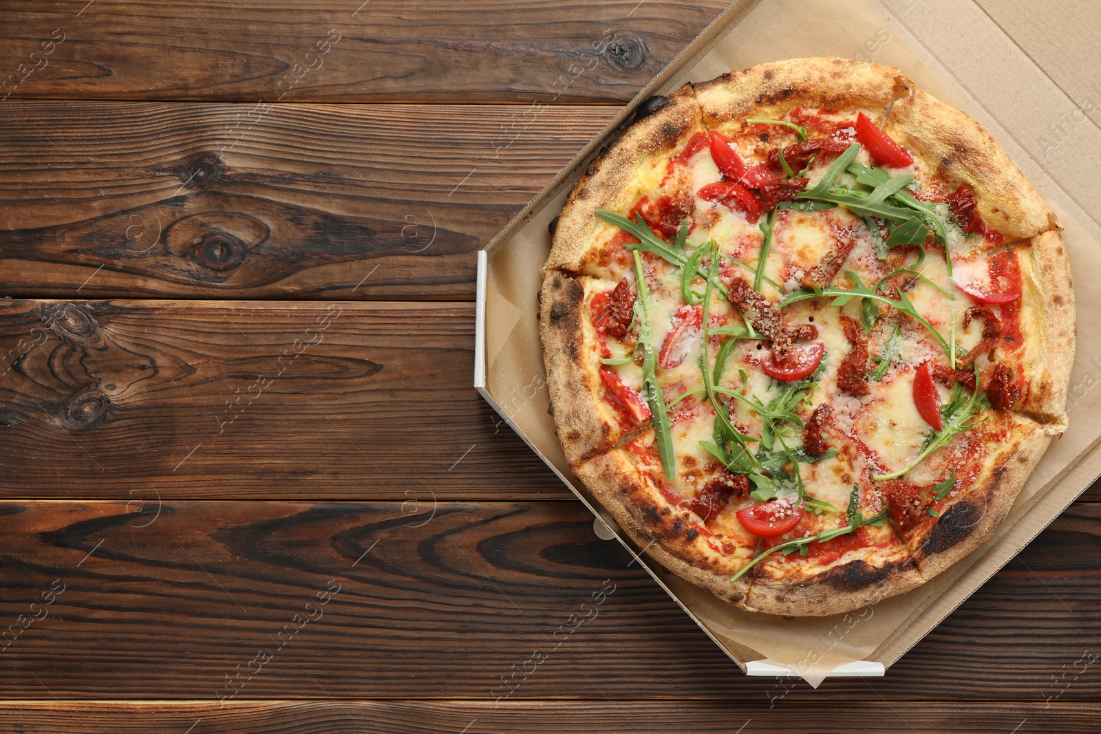 Photo of Tasty pizza with cheese, tomato and arugula in cardboard box on wooden table, top view. Space for text