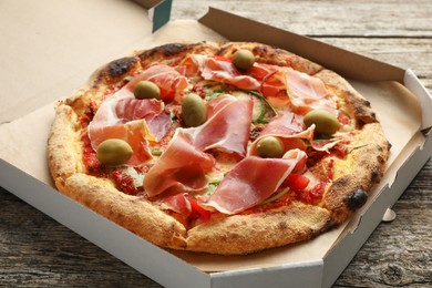 Tasty pizza with cured ham, olives and sun-dried tomato in cardboard box on wooden table