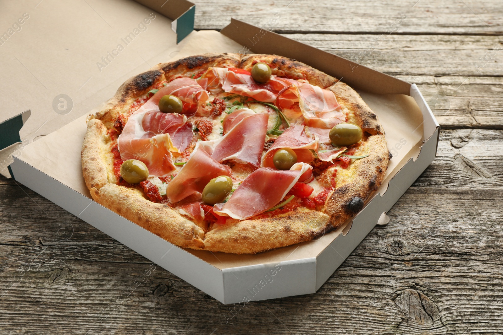 Photo of Tasty pizza with cured ham, olives and sun-dried tomato in cardboard box on wooden table