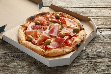 Photo of Tasty pizza with cured ham, olives and sun-dried tomato in cardboard box on wooden table