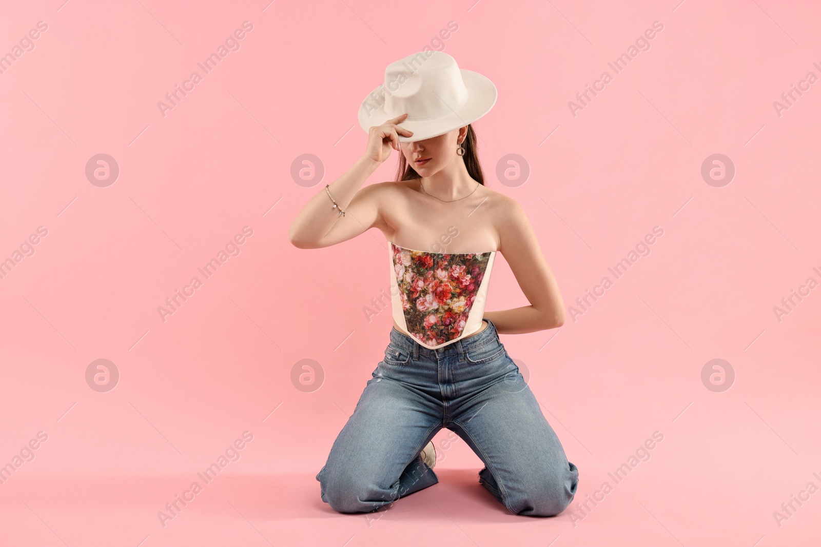 Photo of Woman in stylish corset covering eyes with hat on pink background