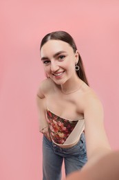 Photo of Smiling woman in stylish corset taking selfie on pink background