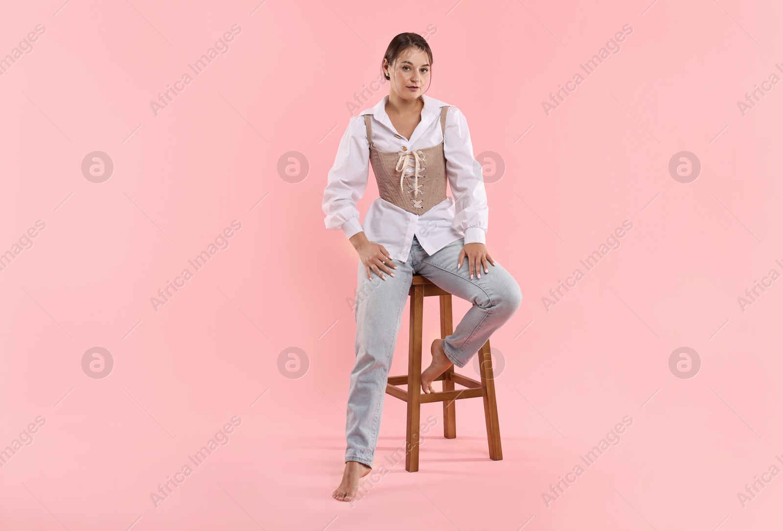 Photo of Beautiful woman in stylish corset sitting on stool against pink background