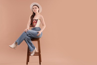 Photo of Beautiful woman in stylish corset and hat posing on stool against beige background. Space for text