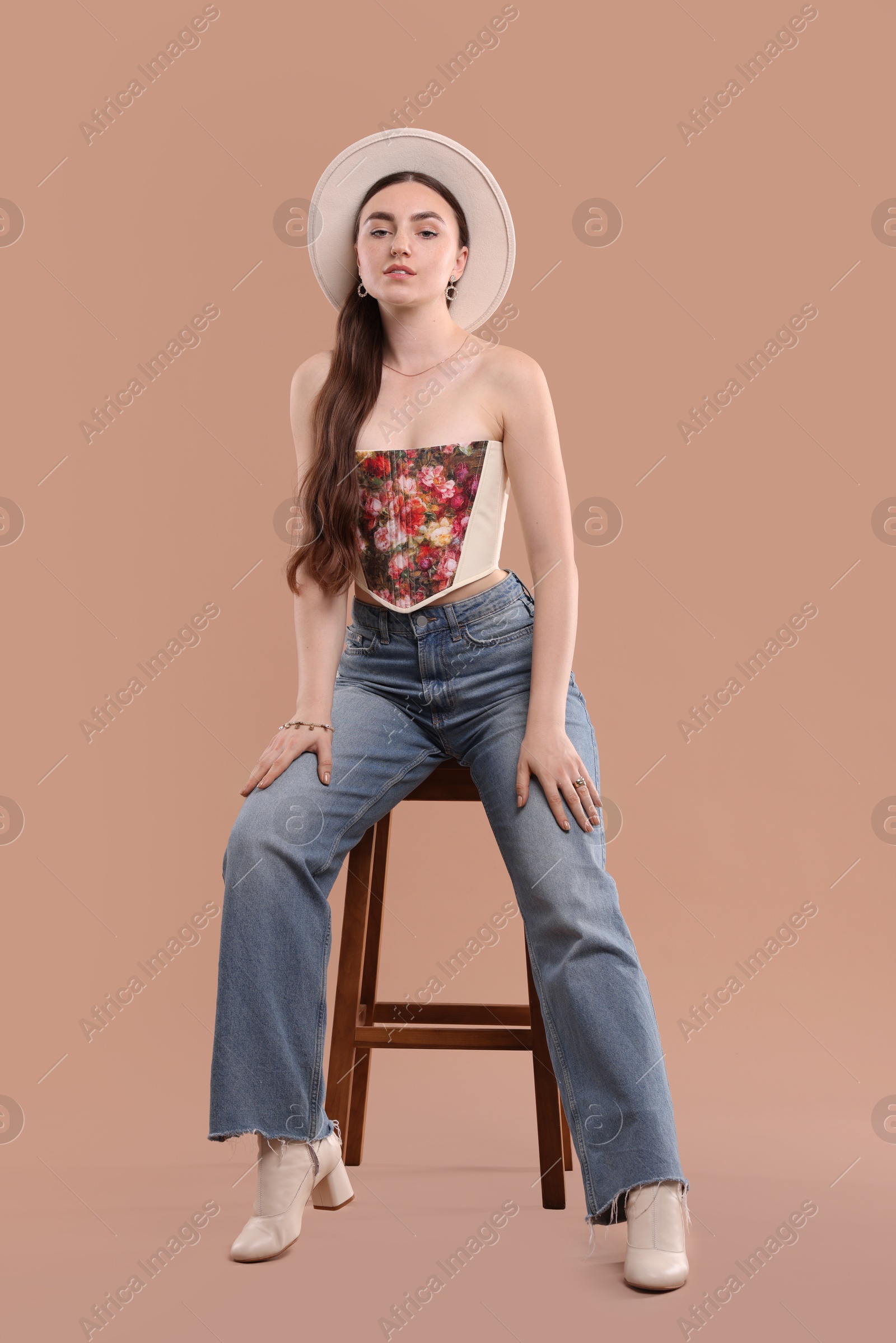 Photo of Beautiful woman in stylish corset and hat posing on stool against beige background