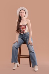 Photo of Beautiful woman in stylish corset and hat posing on stool against beige background