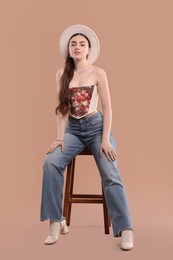 Beautiful woman in stylish corset and hat posing on stool against beige background