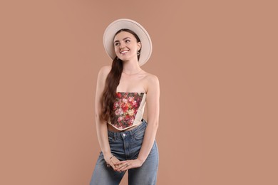 Smiling woman in stylish corset and hat posing on beige background