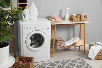 Photo of Washing machine, storage bench, detergents, laundry and houseplant indoors