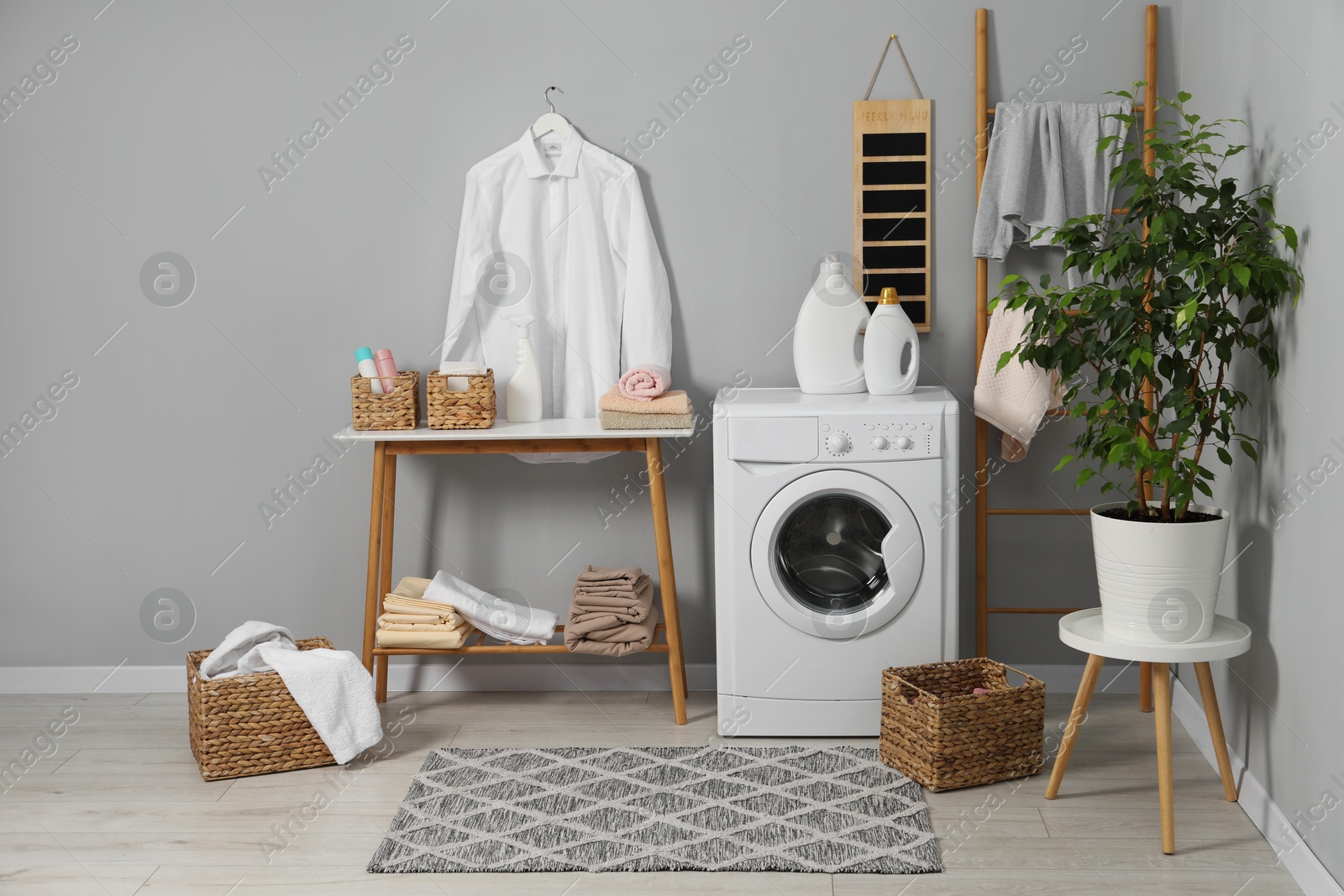 Photo of Washing machine, storage bench, detergents, laundry and houseplant indoors