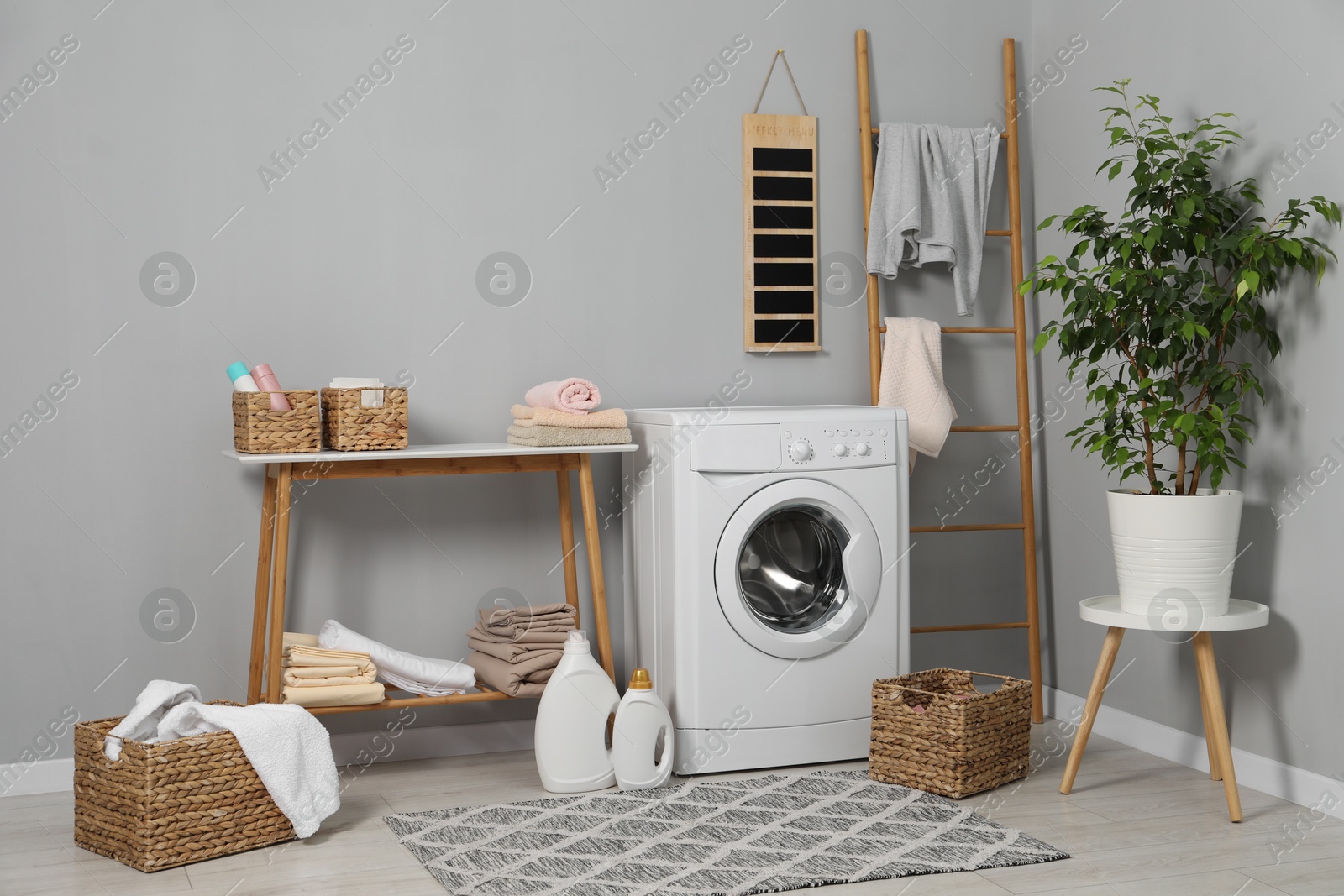 Photo of Washing machine, storage bench, detergents, laundry and houseplant indoors