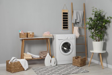 Photo of Washing machine, storage bench, detergents, laundry and houseplant indoors