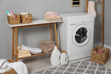 Photo of Washing machine, detergents, basket, laundry and storage bench indoors