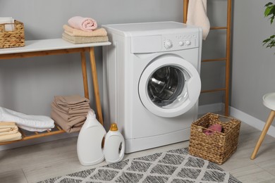 Photo of Washing machine, detergents, basket, laundry and storage bench indoors