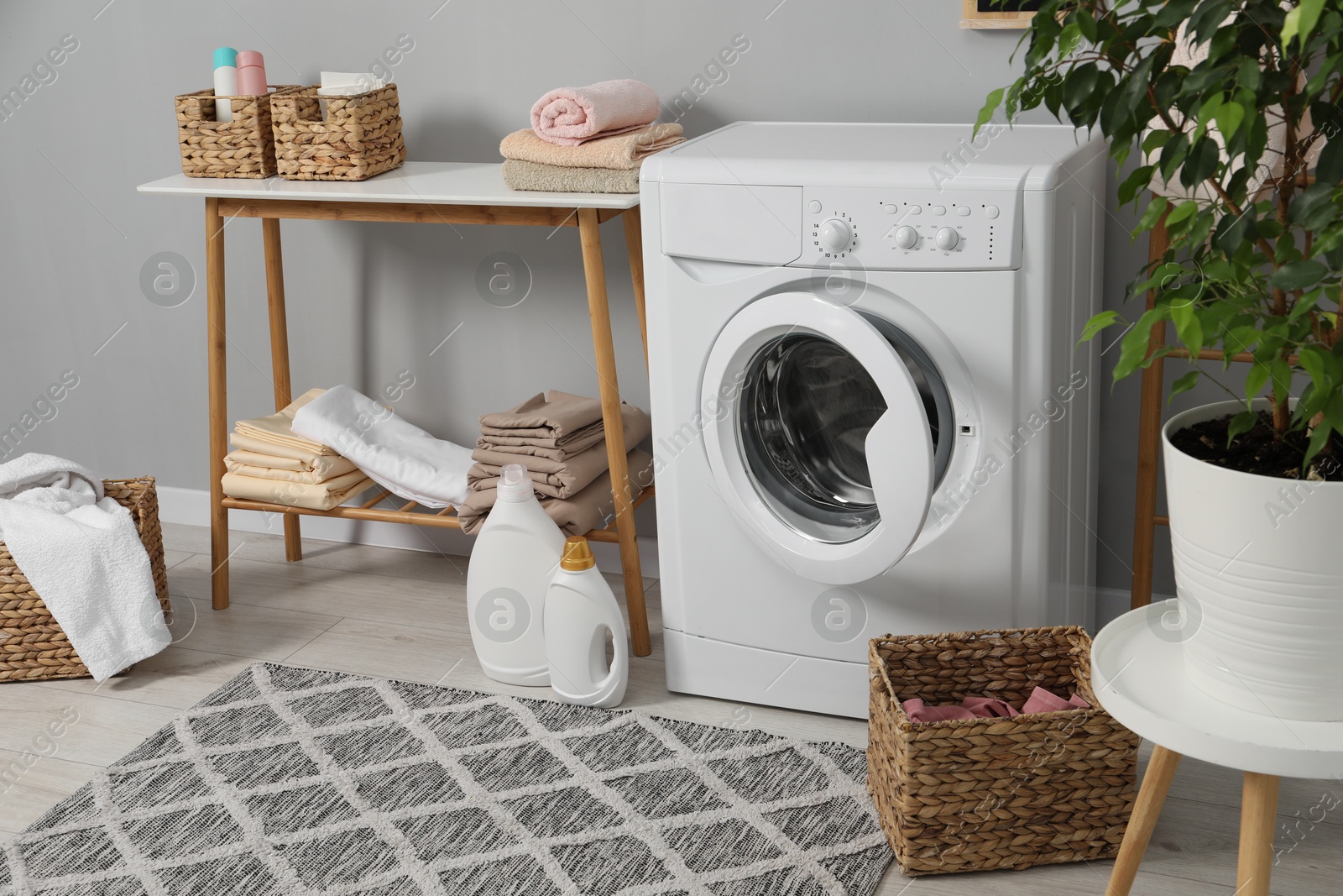 Photo of Washing machine, detergents, basket, laundry, houseplant and storage bench indoors