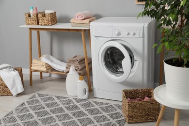 Photo of Washing machine, detergents, basket, laundry, houseplant and storage bench indoors