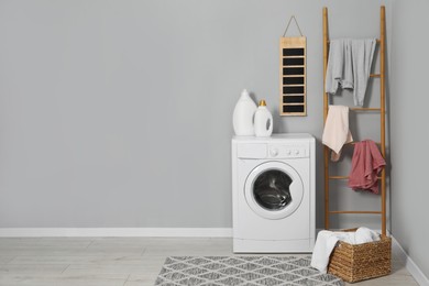 Washing machine, detergents, ladder, basket and laundry near grey wall indoors, space for text