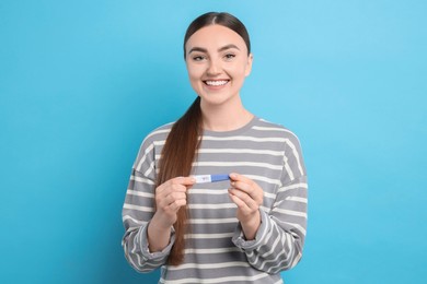 Happy woman holding pregnancy test on light blue background