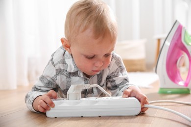 Photo of Little child playing with power strip and iron plug on floor indoors. Dangerous situation