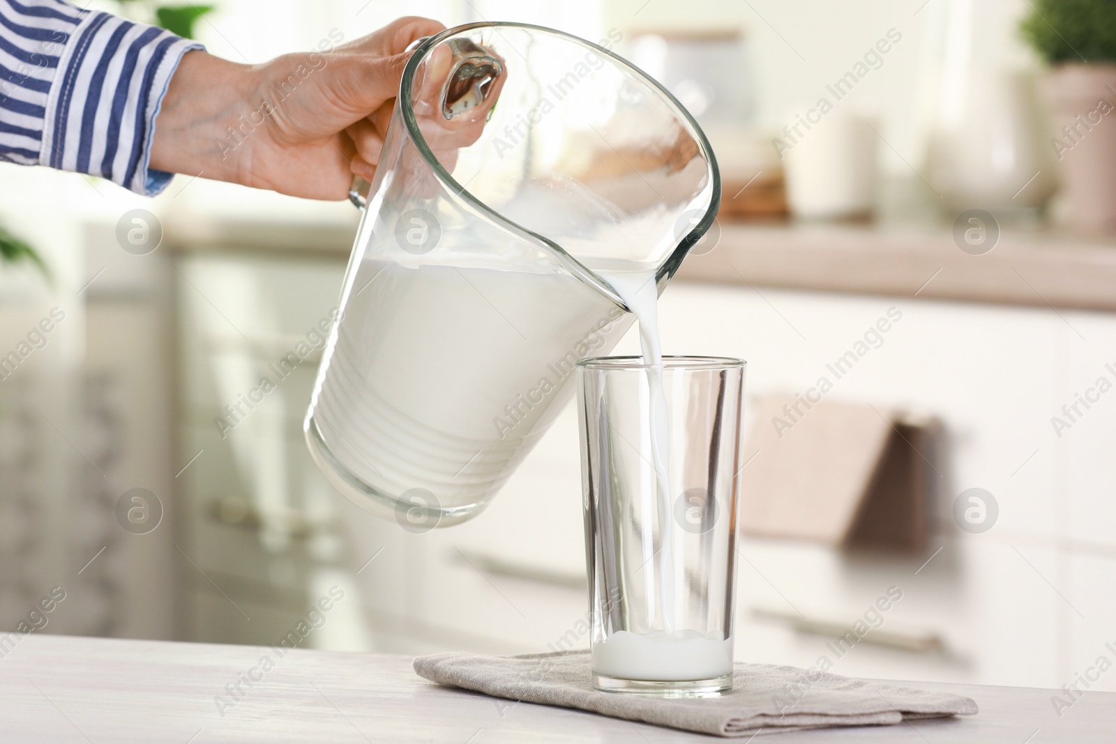 Photo of Woman pouring fresh milk from jug into glass at light wooden table in kitchen, closeup