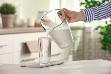 Woman pouring fresh milk from jug into glass at light wooden table in kitchen, closeup