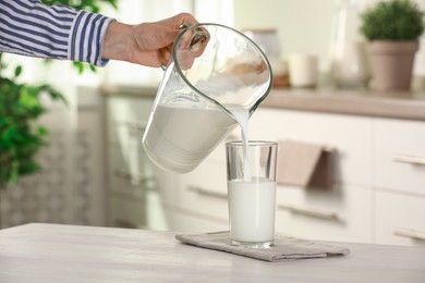 Photo of Woman pouring fresh milk from jug into glass at light wooden table in kitchen, closeup