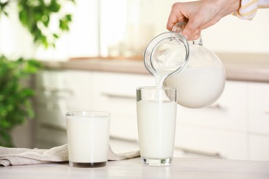 Photo of Woman pouring fresh milk from jug into glass at light wooden table in kitchen, closeup