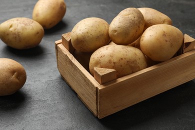 Many fresh potatoes and wooden crate on black textured table, closeup