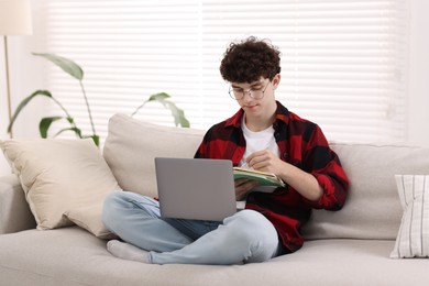 Teenager taking notes while working with laptop on sofa at home. Remote job