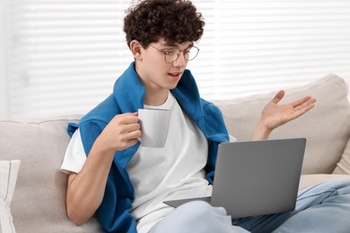 Teenager holding cup of drink working with laptop on sofa at home. Remote job
