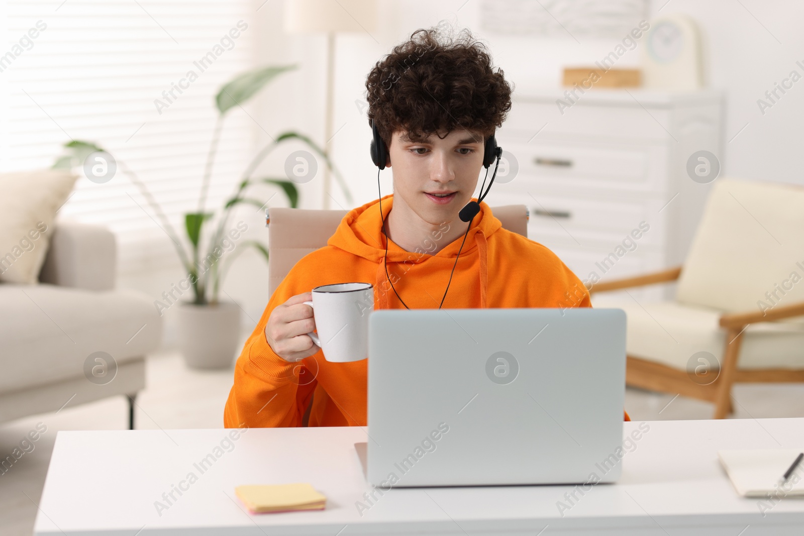 Photo of Teenager in headset holding cup of drink and working with laptop at home. Remote job