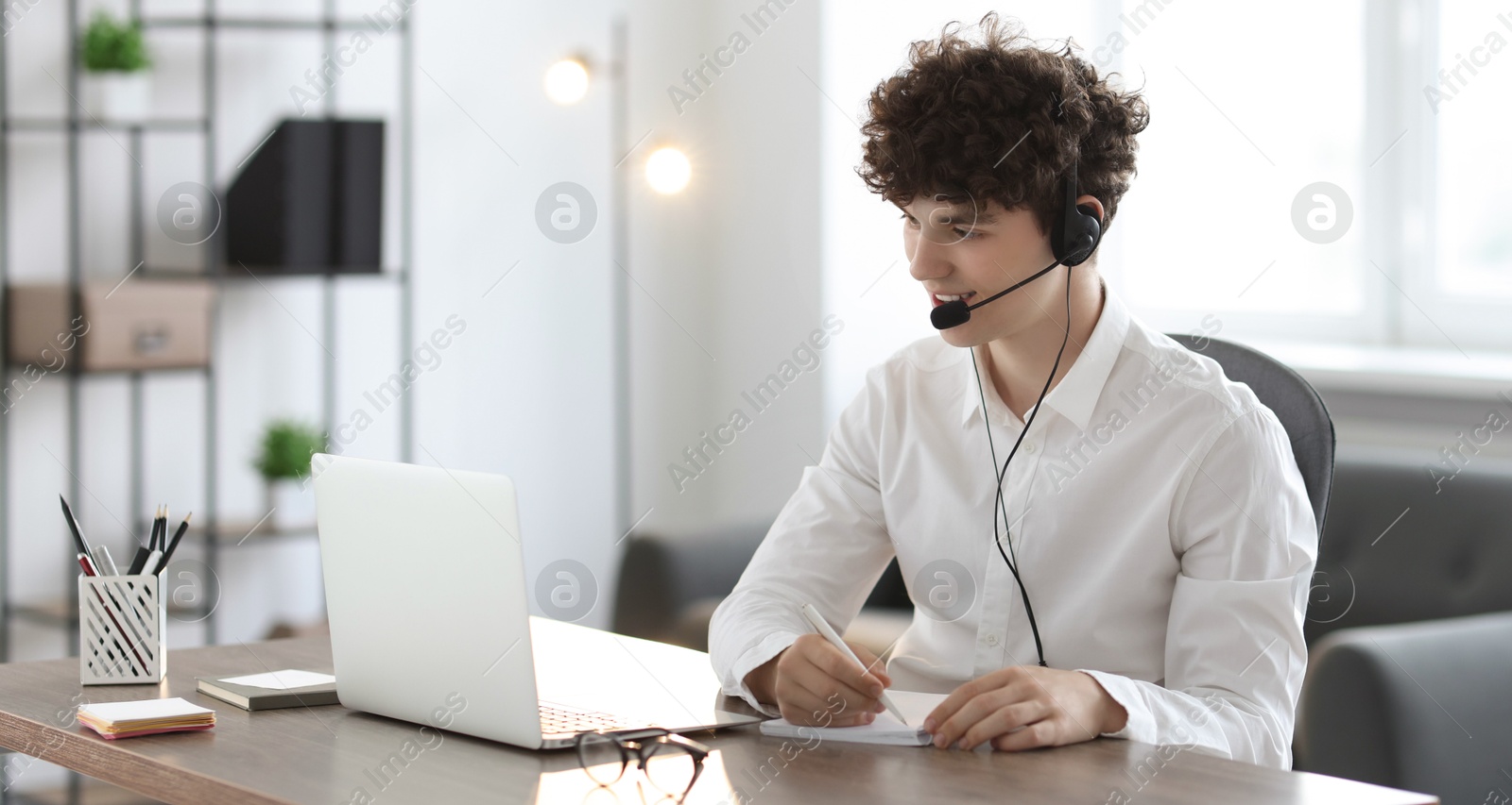 Photo of Teenager in headset taking notes while working with laptop at table indoors. Remote job