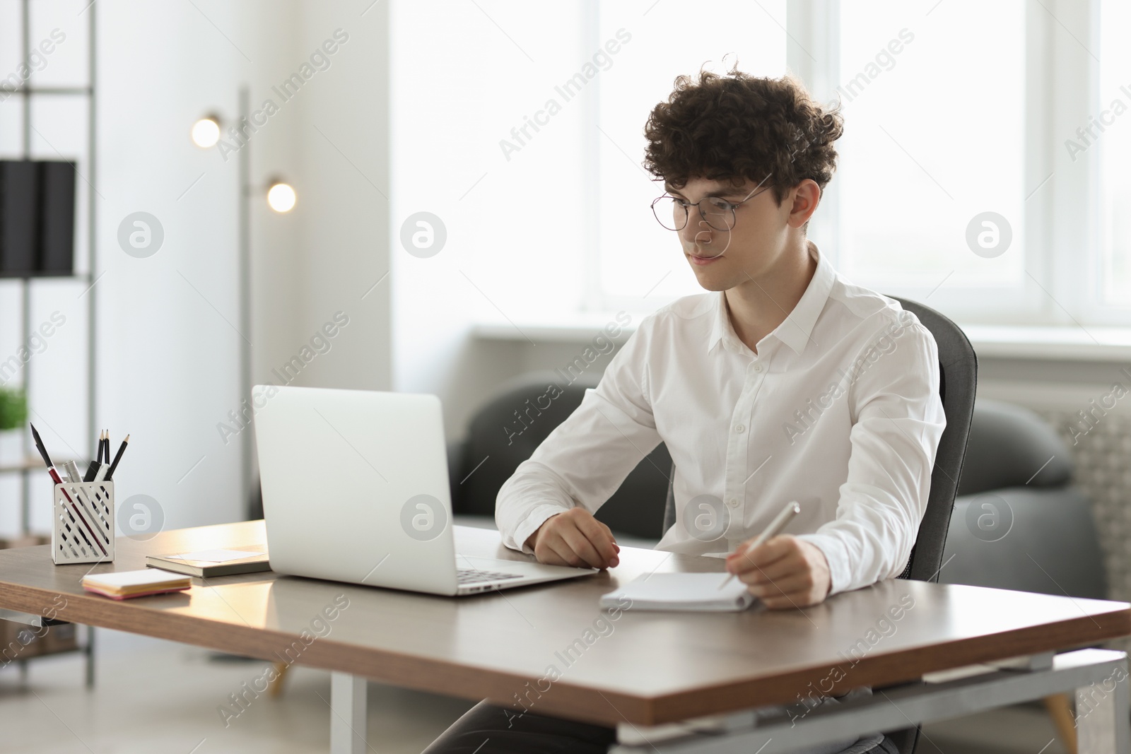 Photo of Teenager taking notes while working with laptop at table indoors. Remote job