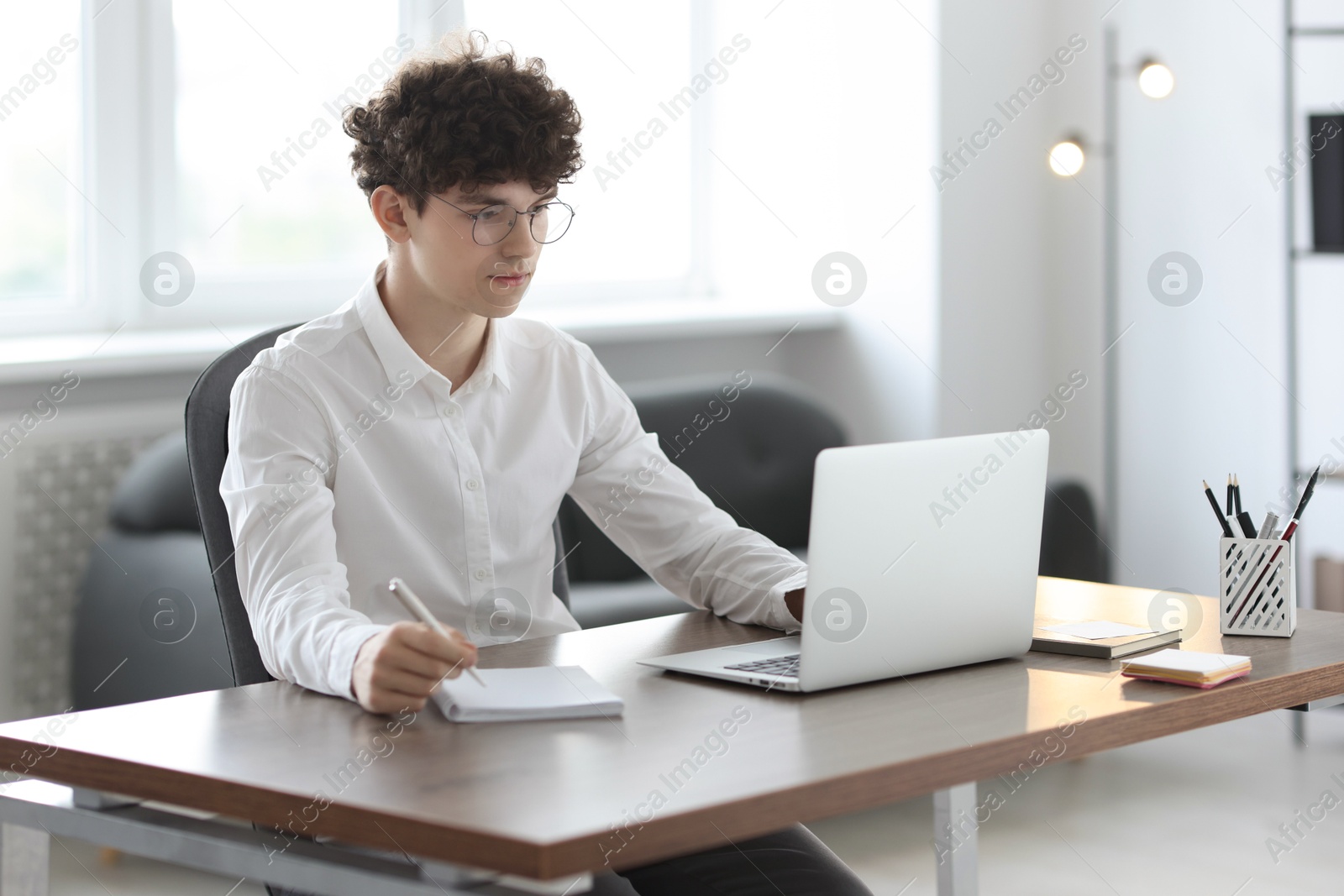 Photo of Teenager taking notes while working with laptop at table indoors. Remote job