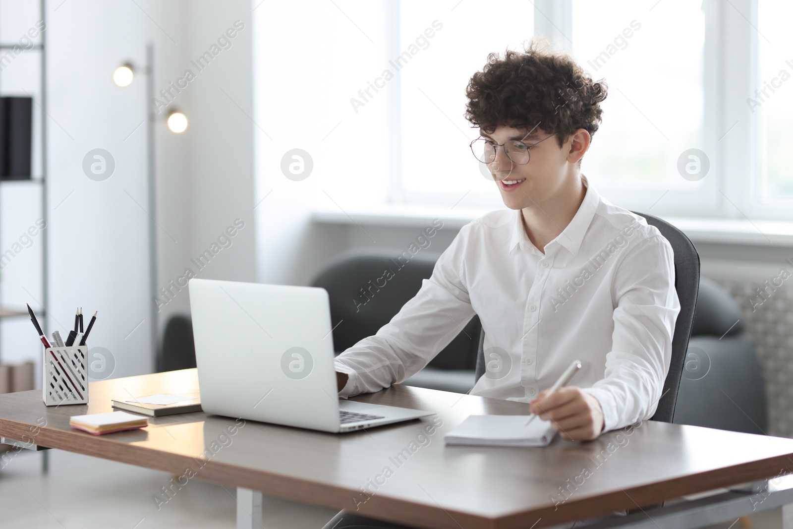 Photo of Teenager taking notes while working with laptop at table indoors. Remote job