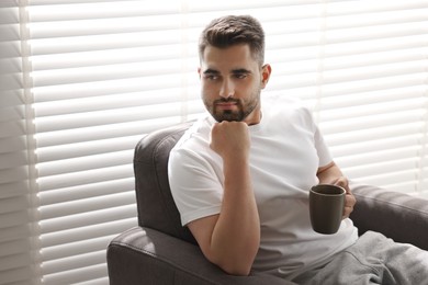 Man with cup of drink sitting on armchair near window blinds indoors