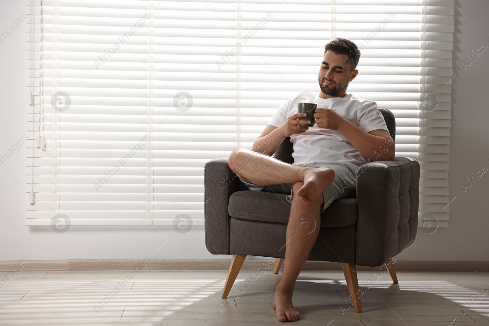 Photo of Man with cup of drink sitting on armchair near window blinds indoors, space for text