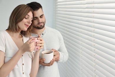 Couple with cups of drink near window blinds at home, space for text