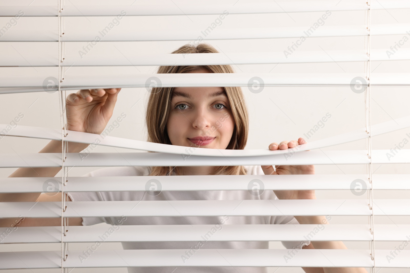 Photo of Young woman looking through window blinds on white background