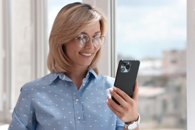 Happy woman using mobile phone near window indoors