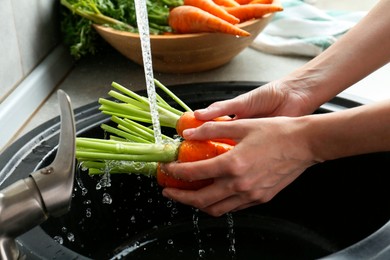 Photo of Woman washing fresh carrots under tap water in above sink indoors, closeup