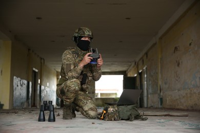 Photo of Military mission. Soldier in uniform with drone controller, laptop and binoculars inside abandoned building