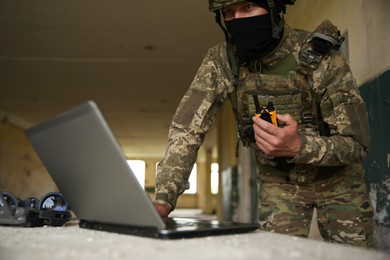 Military mission. Soldier in uniform with radio transmitter using laptop at table inside abandoned building