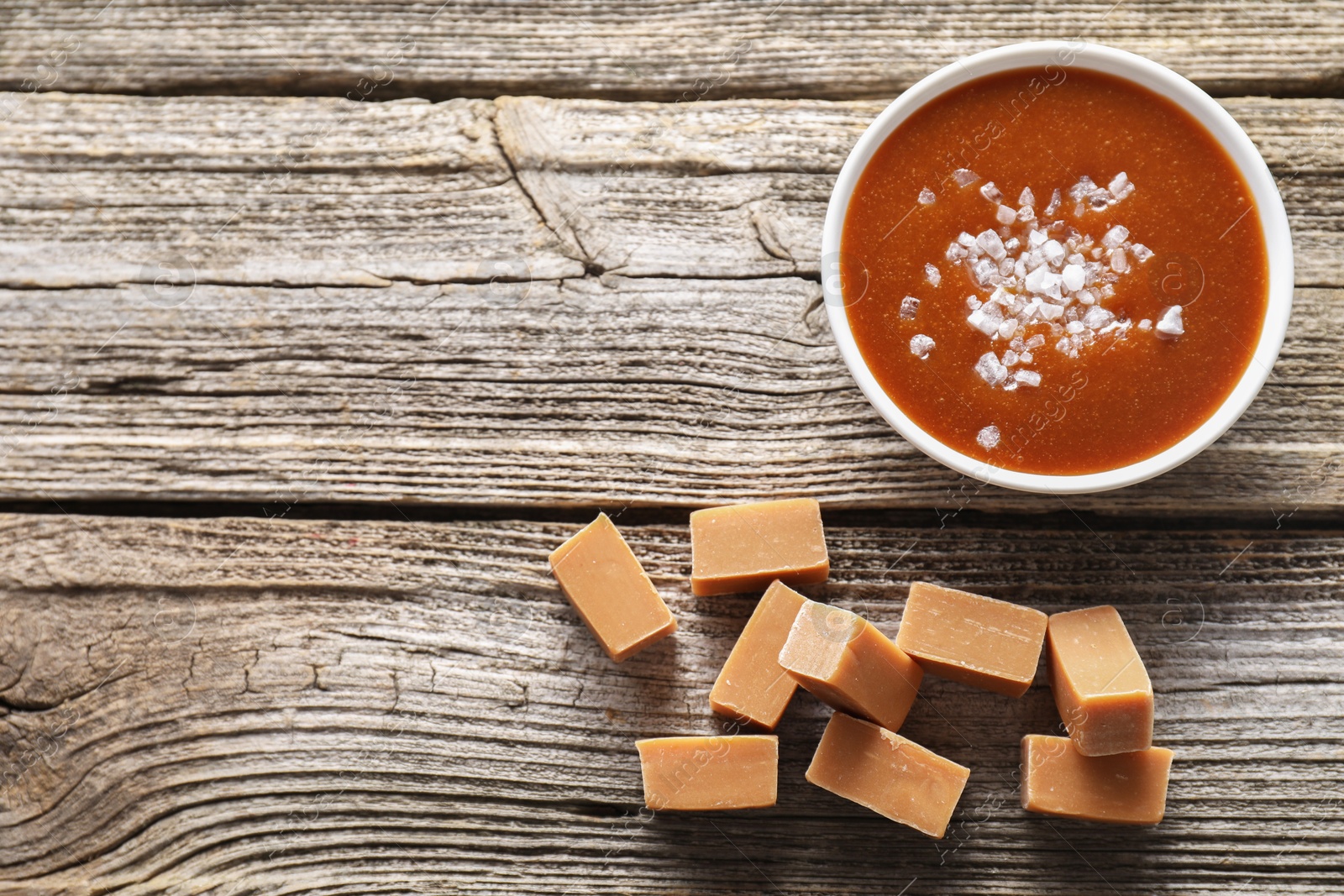 Photo of Caramel sauce with sea salt and yummy candies on wooden table, flat lay. Space for text