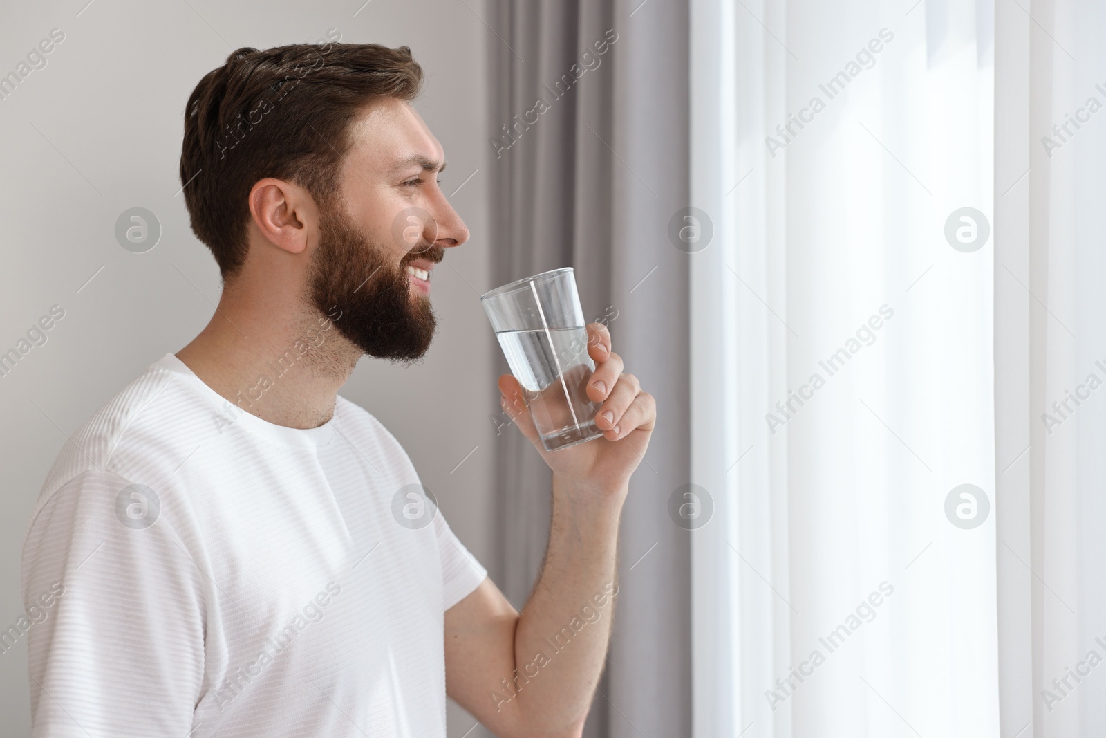 Photo of Happy young man with glass of water near window at morning