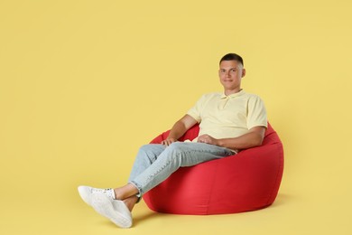 Handsome man sitting on red bean bag chair against yellow background. Space for text