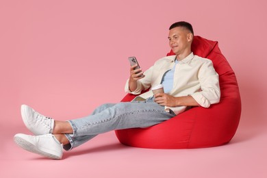 Photo of Happy man with smartphone and paper cup on red bean bag chair against pink background