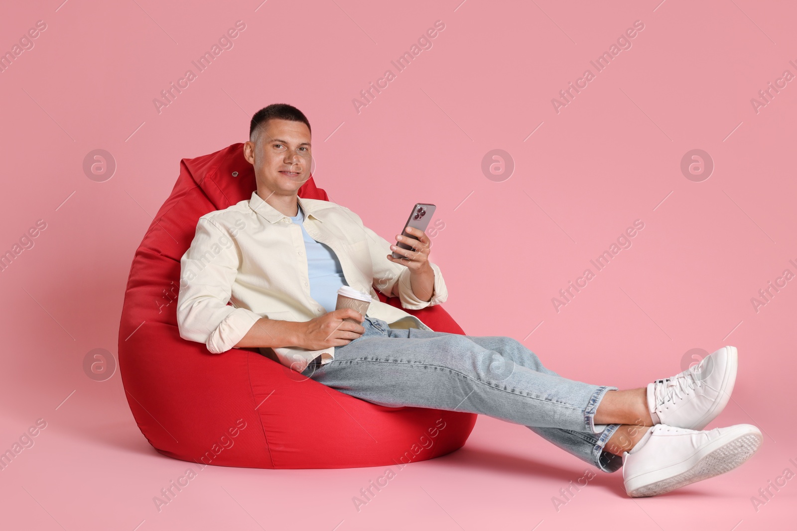 Photo of Happy man with smartphone and paper cup on red bean bag chair against pink background