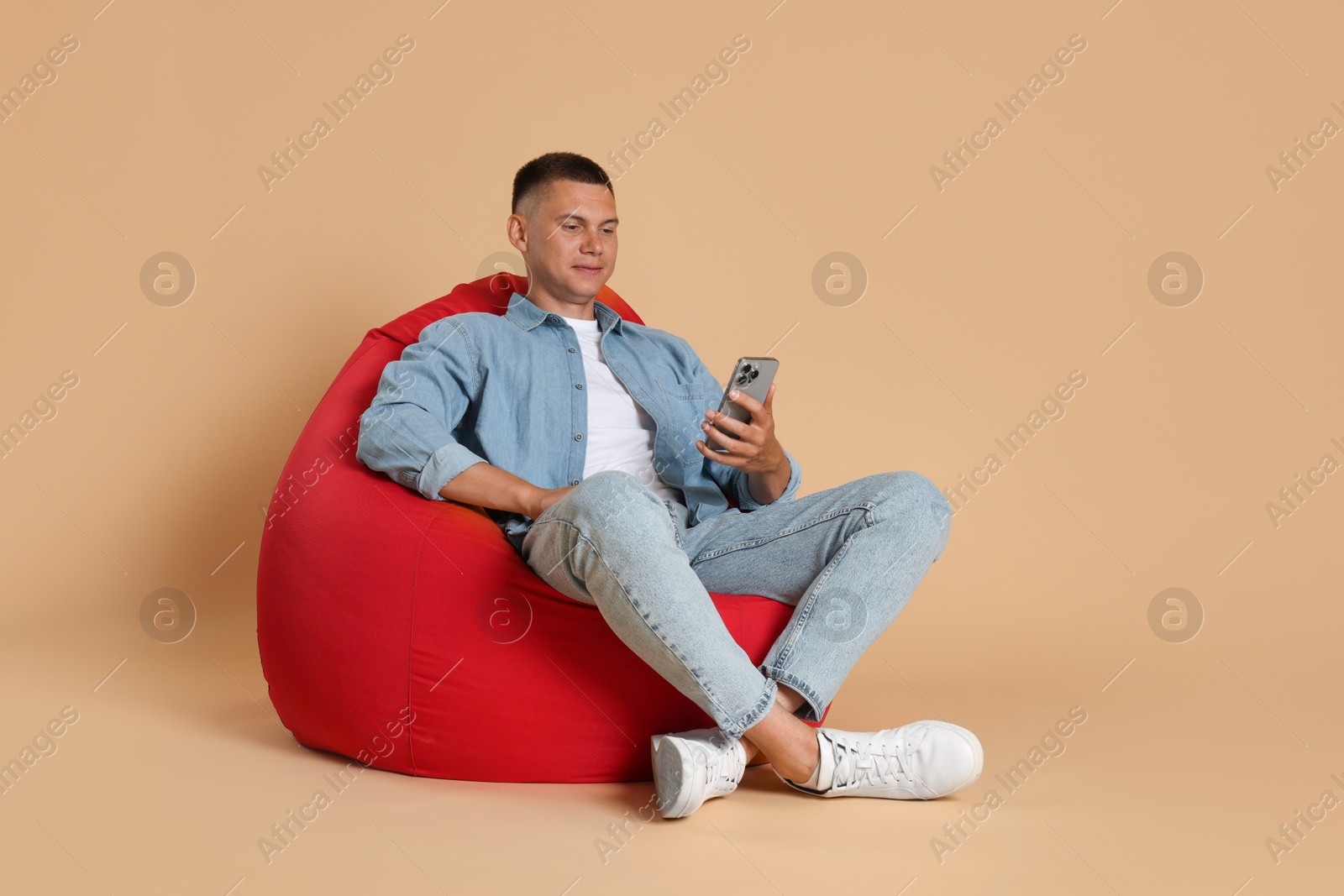 Photo of Handsome man with smartphone on red bean bag chair against beige background