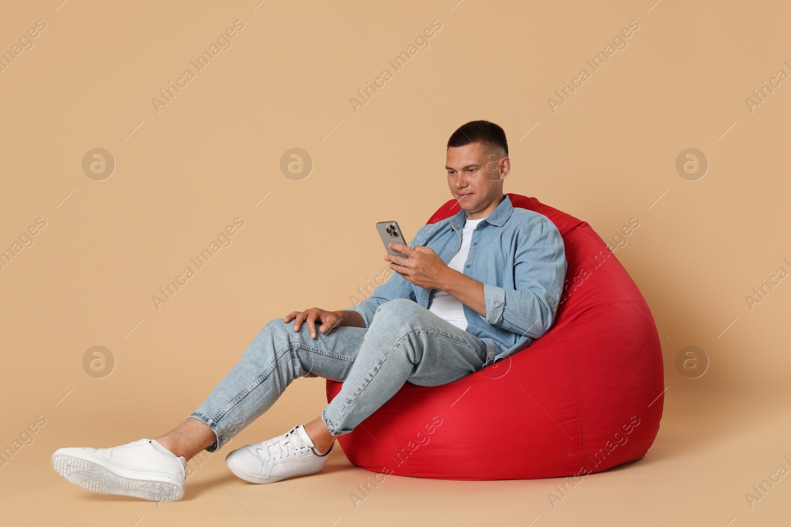 Photo of Handsome man with smartphone on red bean bag chair against beige background